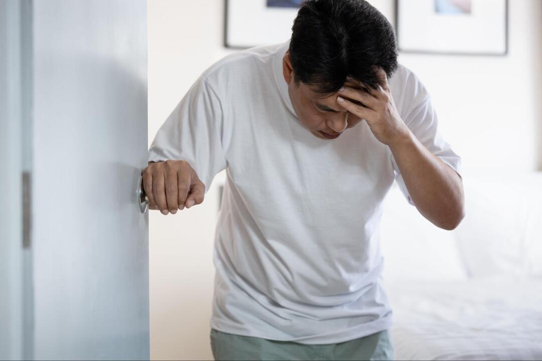 男性が、ドアノブに手をかけながらもう片方の手で額を抑え、苦しそうに下を向いている。