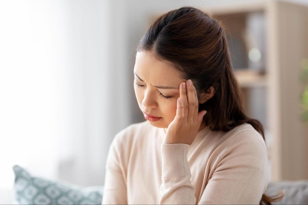女性が、目を閉じて眉間にシワを寄せながら左手でこめかみを抑えている様子