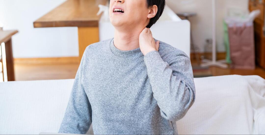 男性が、苦悶の表情を浮かべながら左手で首の後ろ(うなじ辺り)を押さえている様子