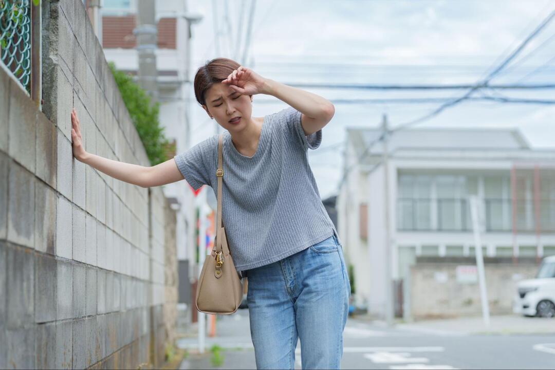 道端のブロック塀に片手を突きながら、女性が額に手を当ててめまいや体調不良に耐えている様子