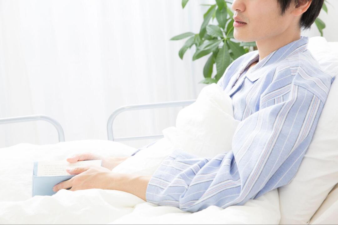 ストライプ柄のパジャマを着た男性が、病室のベッドの上で上半身を起こし、手元で冊子を開いて持っている