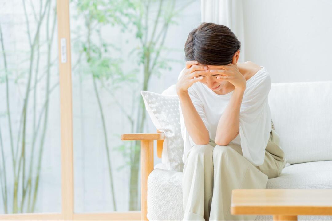 ソファーに腰掛けた女性が、両手を額に当てて下を向き、体調不良や悩みに耐えている様子