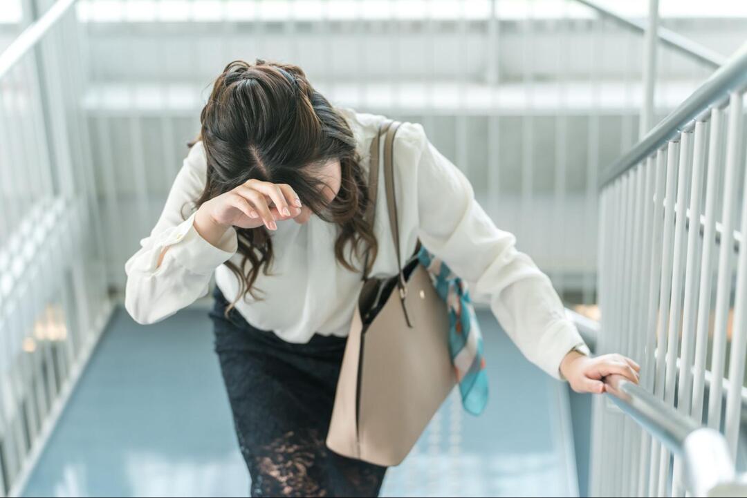 女性が階段を上る途中で手すりを掴みながら、もう片方の手で額を抑えて苦しそうに下を向いている様子