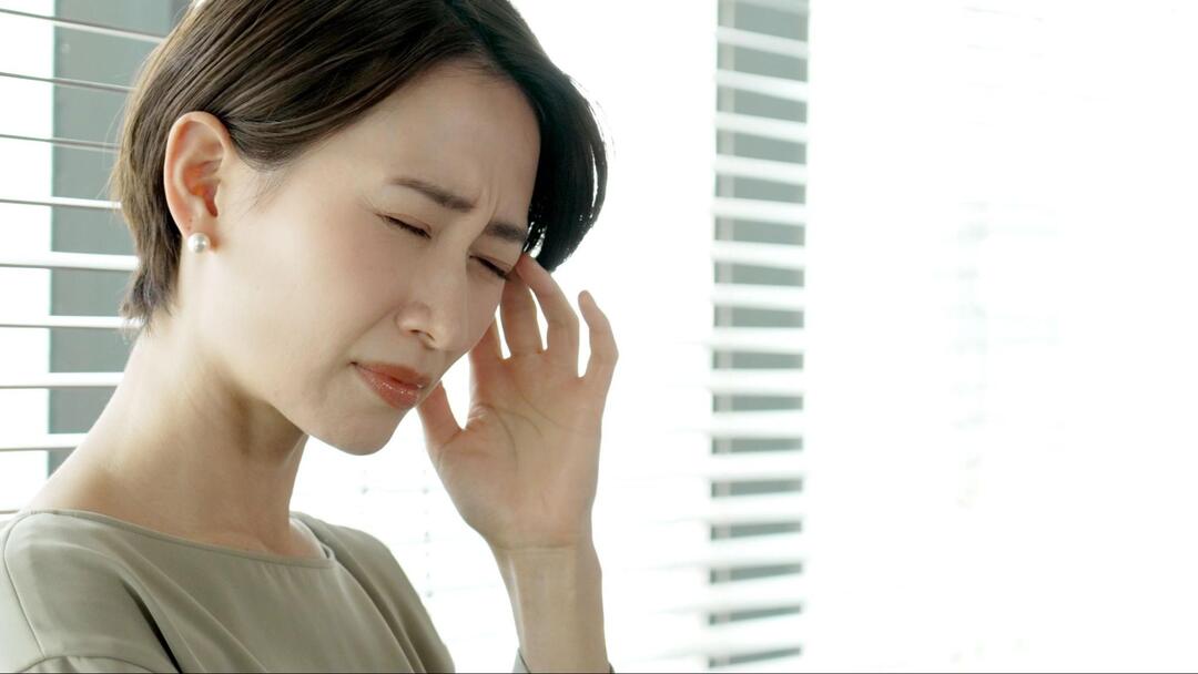 女性が目を閉じながら、左手でこめかみを抑えて頭痛に耐えている表情のアップ