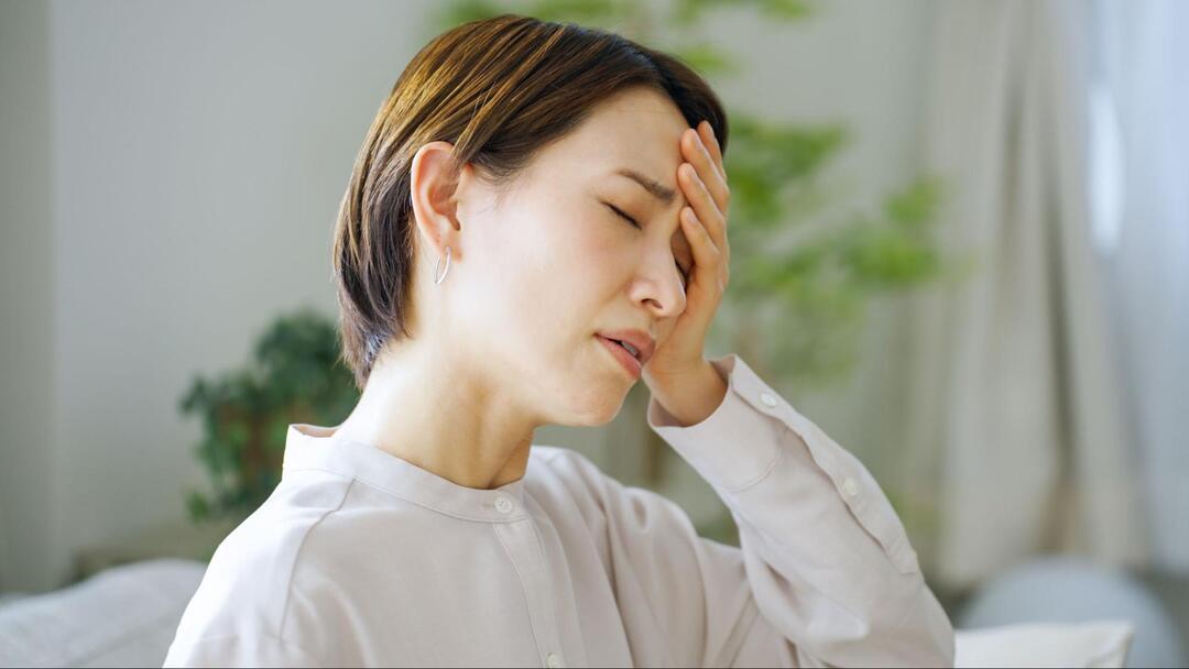 女性が目を閉じながら、左手で額を抑えて頭痛に耐えている様子