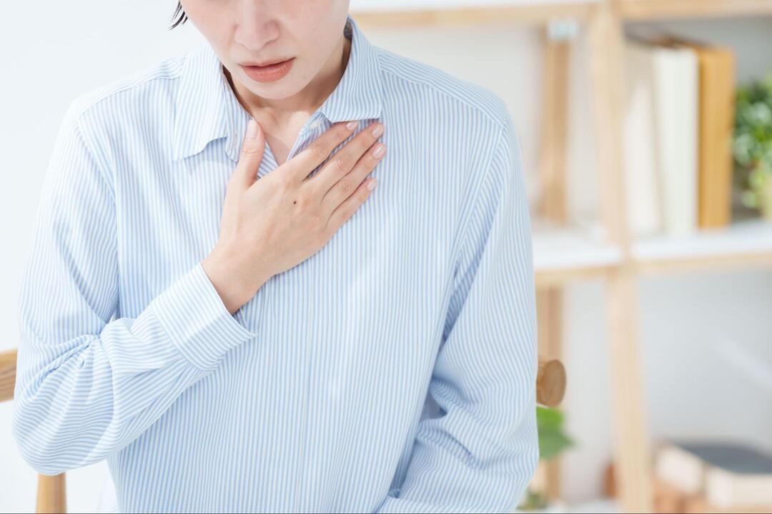水色のストライプ柄のシャツを着た女性が、苦しそうな表情を浮かべながら左手を胸元に当てている様子