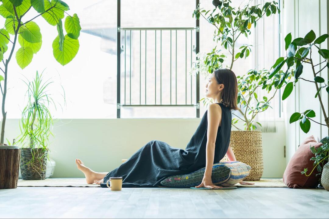 多くの観葉植物が置かれた明るい窓際で、女性が床の上のクッションに座り、リラックスしている様子