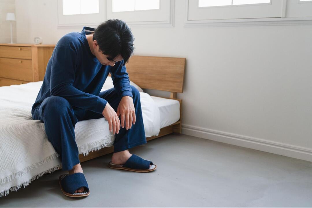 男性がベッドの端に腰掛けて頭を下げ、うつむいている様子