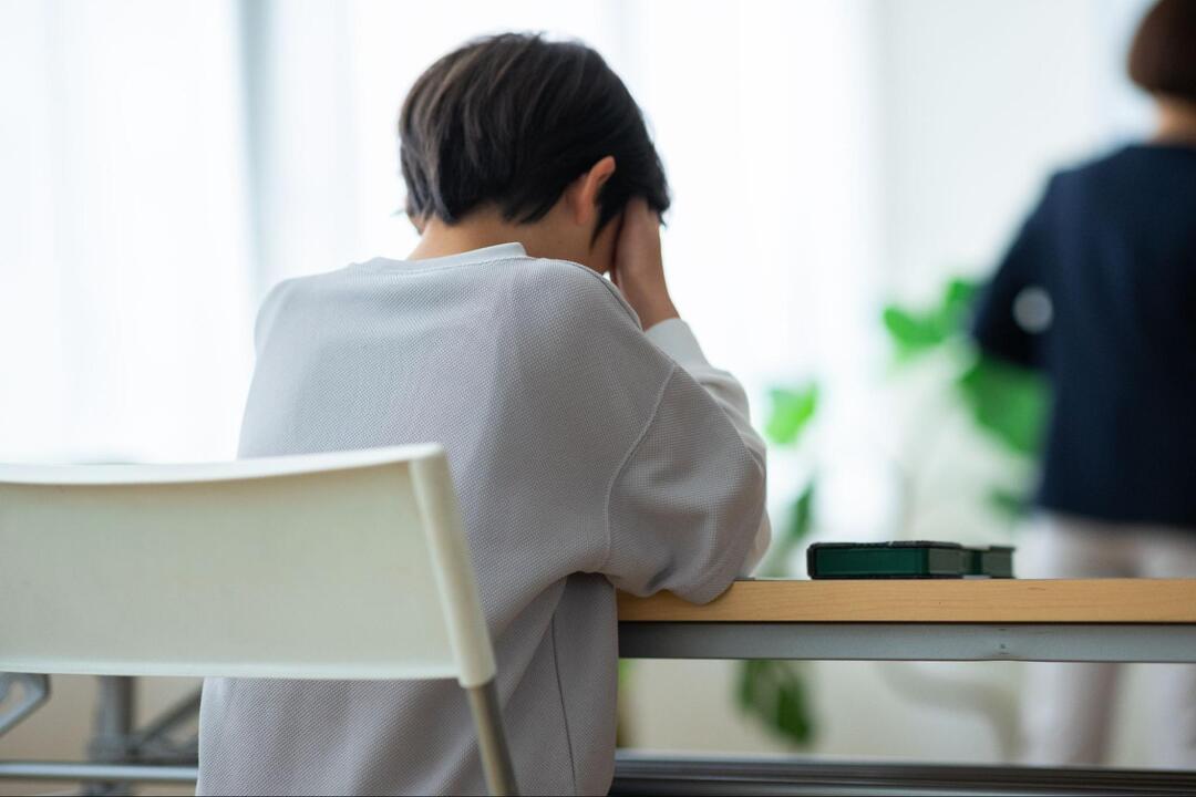 デスクの椅子に座り、両手で頭を抱えてうつむいている人物の後ろ姿