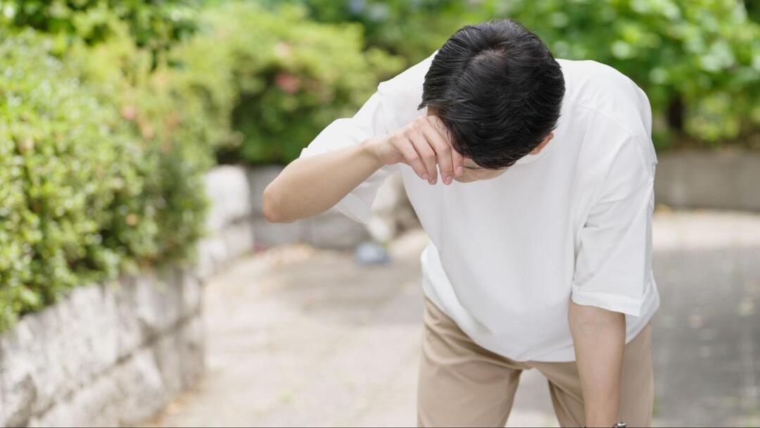 屋外で男性が前かがみになり、片手を額に当てて目眩や頭痛に耐えているような様子